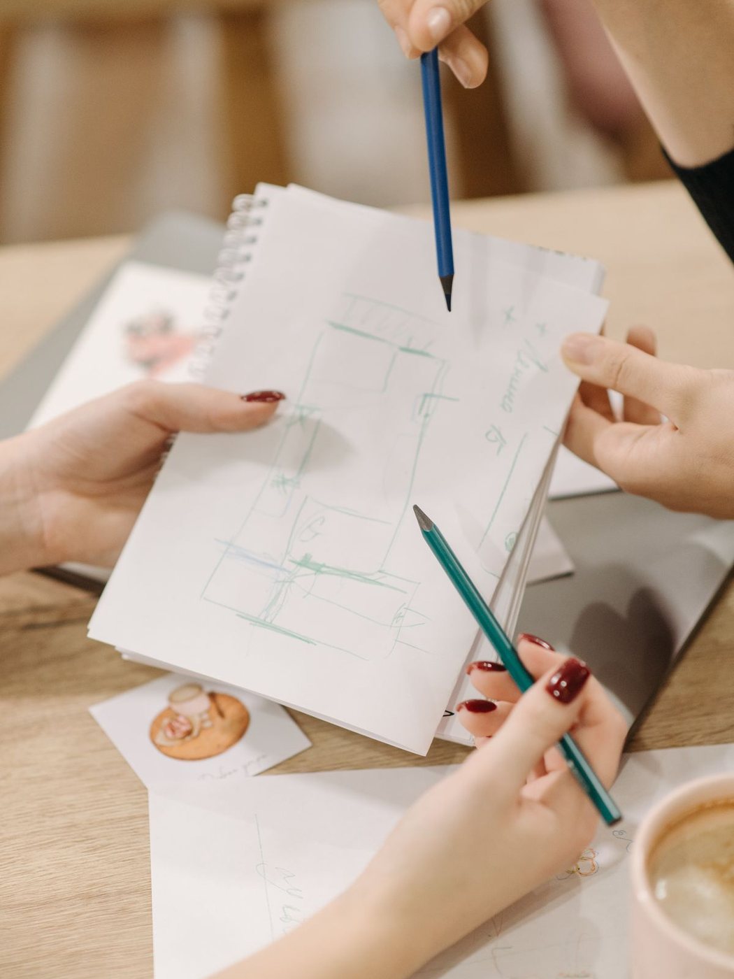 Cuatro manos sobre un cuaderno de bocetos señalando un esquema a lápiz, junto a una taza de café