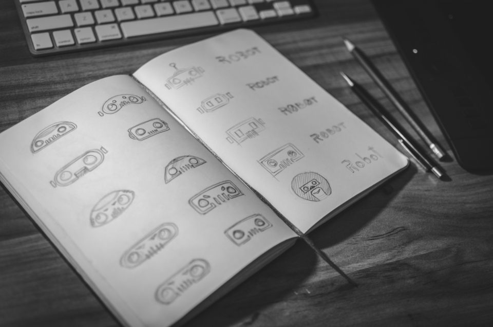 Cuaderno de bocetos abierto con varios conceptos de logotipo dibujados a lápiz, junto a un teclado y dos lapiceros