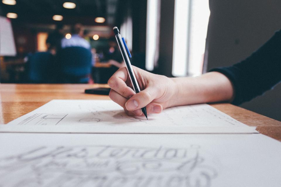 Una mano dibuja a lápiz un rótulo caligráfico sobre un folio, en la mesa de madera de una cafetería